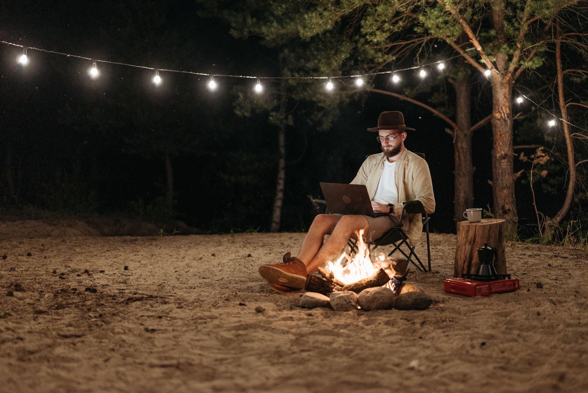 Un freelance installe son ordinateur portable en plein air, près d'un feu de camp, sous une guirlande lumineuse, pour travailler à distance.