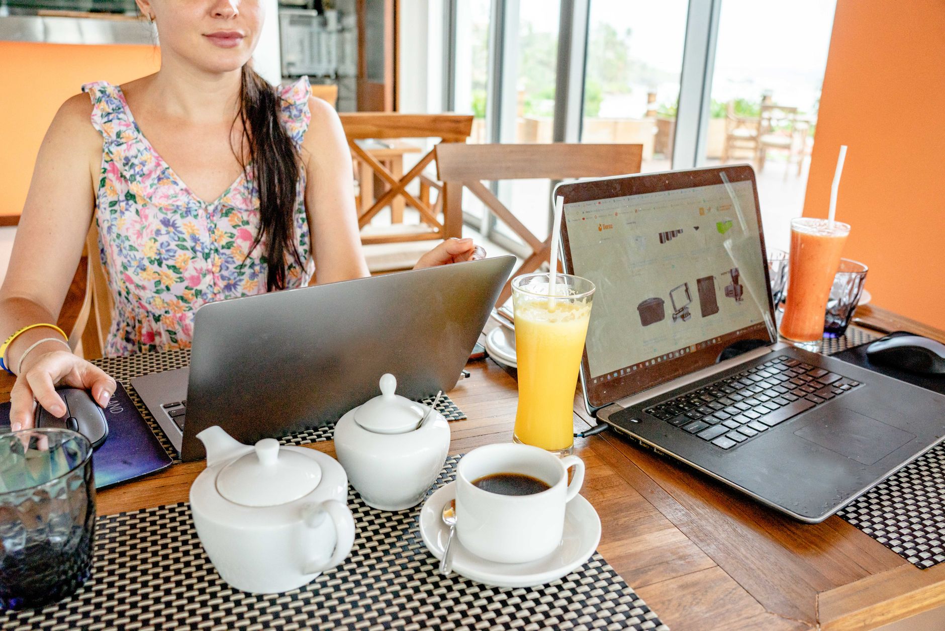 Une femme travaille sur deux ordinateurs portables installée à la table d'un café, boissons chaudes et froides à portée de main.