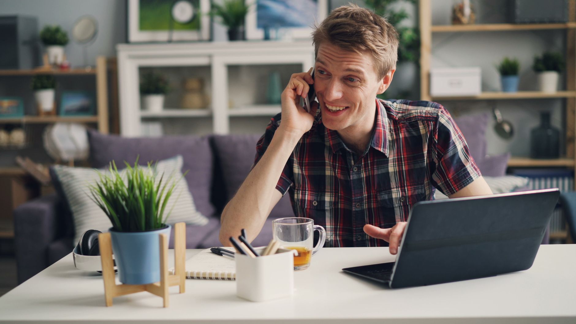 Un homme en chemise à carreaux échange au téléphone avec un client, installé à son bureau à domicile avec un ordinateur portable et un café.